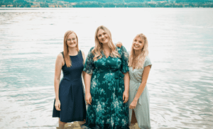 Drei Frauen stehen im seichten Wasser an einem See und diskutieren fröhlich über die Hochzeitsplanung. Sie lächeln und tragen Kleider: eine in Marineblau, eine andere in Blaugrün mit Blumenmuster und die dritte in Hellgrün. Der ruhige See und das entfernte Ufer bieten die perfekte Kulisse für ihre Hochzeitsträume.