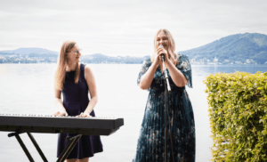 Zwei Frauen treten im Freien an einem ruhigen See auf, der sich perfekt als Hochzeitskulisse eignet. Eine spielt Keyboard, während die andere in ein Mikrofon singt. Ihr freudiges Engagement verleiht dieser idealen Kulisse für jede Hochzeitsplanung Charme. Berge und ruhiges Wasser runden die malerische Szene ab.