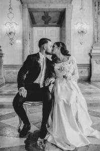Braut und Bräutigam küssen sich in einem eleganten Innenraum und halten damit einen zeitlosen Moment ihrer Hochzeit fest. Die Braut trägt ein langes weißes Kleid und einen Schleier und hält einen Blumenstrauß, während der Bräutigam einen dunklen Anzug trägt. Verzierte Wanddekorationen und Kronleuchter zieren den Hintergrund. Schwarzweißbild.