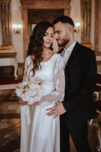 In einem eleganten Raum mit kunstvollem Dekor posiert ein Brautpaar in zeitloser Harmonie. Bei der Feier ihrer Hochzeit trägt die Braut ein weißes Kleid mit Spitzenärmeln und hält einen Strauß weißer und beiger Blumen. Der Bräutigam ergänzt ihre Schönheit in einem schwarzen Anzug und hält seine Geliebte sanft im Arm.