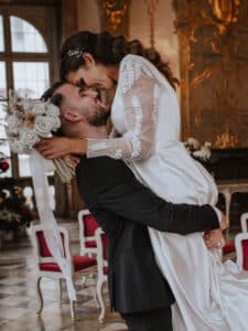 Ein Bräutigam hebt seine Braut in einer freudigen Umarmung hoch und feiert ihre Hochzeit. Die Braut hält einen Strauß weißer Blumen und trägt ein elegantes weißes Kleid mit Spitzenärmeln. Sie befinden sich in einem großen, kunstvoll dekorierten Raum mit großen Fenstern und roten Stühlen.