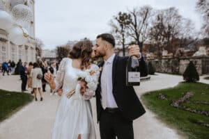 Ein frisch vermähltes Paar küsst sich draußen in einem Park. Die Braut umklammert weiße Luftballons und einen Strauß Rosen, während der Bräutigam eine Champagnerflasche hält. Elegant gekleidet inmitten ihrer Hochzeitsfeier teilen sie einen glückseligen Moment, während sich im Hintergrund die Gäste unterhalten.