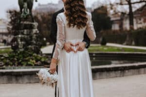 Ein Paar umarmt sich vor einem Brunnen und feiert seine Hochzeit. Die Frau im weißen Kleid hält einen Strauß weißer Rosen und Pampasgras. Der Mann im dunklen Anzug formt mit seinen Händen ein Herz auf ihrem Rücken. Es ist eine Szene im Freien mit unscharfem Hintergrund.