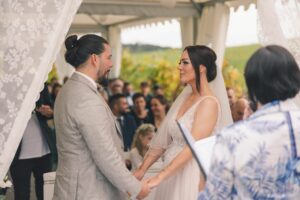 Braut und Bräutigam stehen sich bei einer Hochzeit im Freien gegenüber und halten Händchen. Die Kulisse ist mit durchsichtigen weißen Vorhängen geschmückt und die Gäste sitzen im Hintergrund. Der Bräutigam trägt einen hellen Anzug, während die Braut in ihrem weißen Kleid glänzt.