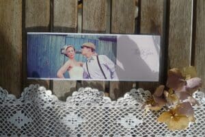 Eine Dankeskarte mit einem Foto im Vintage-Stil eines Paares in Retro-Kleidung, perfekt zur Erinnerung an Ihre besondere Hochzeit. Die Karte liegt auf einer mit Spitze und Trockenblumen geschmückten Holzoberfläche und fängt die Essenz zeitloser Romantik als Teil Ihrer Hochzeitserinnerungen ein.