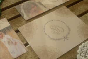 Eine Hochzeitseinladung auf einer Holzoberfläche weckt den Charme eleganter Hochzeitsplanung. Die Hauptkarte zeigt ein elegantes Monogramm und kunstvolle Blumenmuster. Zwei Fotokarten sind teilweise sichtbar und zeigen eine Braut in einem Spitzenkleid und mit Blumenkrone, bereit, die Freude des Heiratens zu genießen.