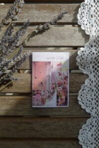 Eine „Save the Date“-Karte mit Blumenmustern liegt auf einem Holztisch neben Lavendelzweigen und einem Spitzendeckchen und schafft die Kulisse für eine elegante Hochzeit. Sonnenlicht schafft eine sanfte, warme Atmosphäre, perfekt, um die Essenz von Heiraten einzufangen.