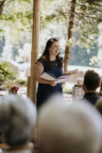 Eine Frau steht im Freien, spricht in ein Mikrofon und hält ein Notizbuch in der Hand. Sie scheint eine Ansprache zu Hochzeitsplanung zu halten, im Vordergrund sind die Teilnehmer verschwommen zu sehen. Sonnenlicht fällt durch die Bäume und schafft eine ruhige Atmosphäre für diesen Hochzeitsanlass.
