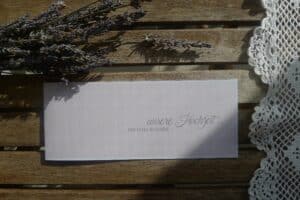 Eine Hochzeitseinladungskarte auf einem Holztisch mit Lavendelzweigen und einer Tischdecke aus Häkelspitze. Auf der Karte steht in eleganter Handschrift „unsere Hochzeit ... Michael & Marie“ und verkörpert damit die Essenz einer durchdachten Hochzeitsplanung.