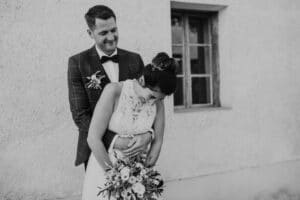 Ein Schwarzweißfoto zeigt ein Paar, das sich vor einem Gebäude mit einem Fenster umarmt und den Geist ihrer Hochzeit verkörpert. Die Frau hält einen Blumenstrauß und lehnt sich nach vorne, während der Mann hinter ihr lächelt und die Arme um ihre Taille geschlungen hat. Beide sind für diesen besonderen Moment elegant gekleidet.