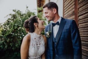 Braut und Bräutigam stehen eng beieinander und lächeln sich an ihrem Hochzeitstag freudig an. Die Braut trägt ein weißes Spitzenkleid und der Bräutigam einen blau karierten Anzug mit Fliege. Sie stehen draußen mit viel Grün und einer Holzwand im Hintergrund und markieren einen Moment perfekter Hochzeitsplanung.