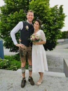 Ein Paar steht an seinem Hochzeitstag zusammen auf einer Steintreppe und lächelt. Der Mann trägt ein traditionelles Outfit mit Weste und Lederhose, während die Frau in ihrem weißen Kleid einen Blumenstrauß hält. Ein grüner Baum bietet die perfekte Kulisse für diesen Moment des Heiratsglücks.
