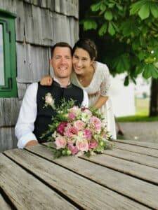 Ein lächelndes Paar posiert im Freien vor einer rustikalen Holzwand und fängt einen Moment purer Freude bei der Hochzeitsplanung ein. Der Mann sitzt mit einem Strauß rosa und weißer Blumen vor ihm auf dem Tisch, während die Frau über seine Schulter gebeugt ist. Üppige grüne Blätter umgeben sie und unterstreichen ihre Liebesfeier.