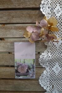 Ein Holztisch mit Spitzendecke, Trockenblumen und einer Karte bildet die Kulisse für die Hochzeitsplanung. Die Karte, geschmückt mit dem Text „Save the Date“ über einem Foto von rosa Blumen in einer silbernen Vase, flüstert das Versprechen einer unvergesslichen Hochzeit.
