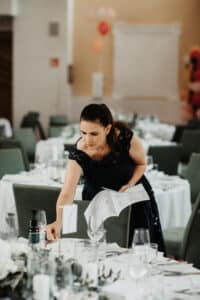 Eine Frau im Abendkleid, vertieft in die Hochzeitsplanung, arrangiert die Tischdekoration an einem dekorierten Veranstaltungsort. Sie hält ein Blatt Papier in der Hand und ordnet Gegenstände auf einem weiß gedeckten Tisch, der von Stühlen umgeben ist. Elegante und ordentlich gedeckte Tische füllen den Raum und schaffen eine anspruchsvolle Atmosphäre für diese Hochzeit.