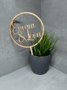 Ein Holzschild mit den Namen „Emma & Leon“ in Schreibschrift steht in einem grauen Topf mit üppigen grünen Pflanzen, perfekt für jede Hochzeit, vor einem eleganten grauen Fliesenhintergrund.