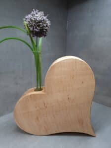 Eine einfache Holzvase in Form eines runden Herzens hält einen einzelnen hohen grünen Stiel mit einer kugelförmigen lila Blüte, perfekt, um Ihrer Hochzeitsdekoration einen Hauch von Eleganz zu verleihen. Der Hintergrund ist eine schlichte, glatte graue Wand.