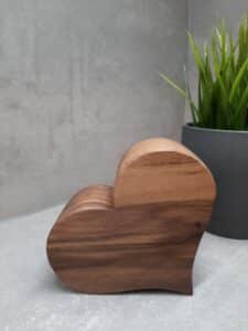 Ein herzförmiges Holzobjekt steht auf einer grauen Oberfläche neben einer grünen Topfpflanze und verleiht einer Hochzeitsplanung Charme. Die strukturierte graue Wand im Hintergrund unterstreicht die einfache und ruhige Umgebung, ideal für diejenigen, die planen, den Bund fürs Leben zu schließen.