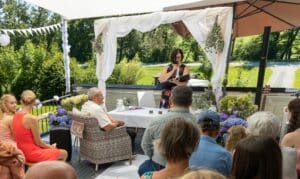 Eine Zeremonie im Freien an einem sonnigen Tag mit einem weißen Baldachin bildet die Kulisse für diese Hochzeit. Eine Frau steht an einem Podium und hält eine Rede, umgeben von sitzenden Gästen. Zur Dekoration gehören Blumen und herabhängendes Laub. Ein Stuhl mit der Aufschrift „Opa“ ist zu sehen und spiegelt eine durchdachte Hochzeitsplanung wider.