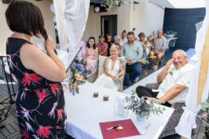 Inmitten einer traumhaften Kulisse im Freien sitzen die Braut in ihrem eleganten weißen Kleid und der Bräutigam in seinem frischen weißen Hemd auf Korbstühlen. Als Teil einer sorgfältigen Hochzeitsplanung spricht eine Frau in einem geblümten Kleid mit einem Mikrofon zu den Gästen, umgeben von leuchtenden Blumen und ätherischen weißen Vorhängen.