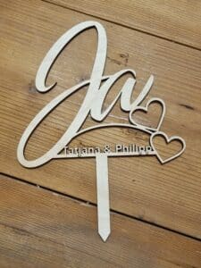 Ein hölzerner Tortenaufsatz auf einer Holzoberfläche, perfekt für eine Hochzeitsfeier. Er hat die Form des Wortes „Ja“ mit zwei Herzumrissen, darunter steht elegant in kleineren Buchstaben „Tatyana & Philipp“, was die Essenz von Heiraten wunderschön einfängt.