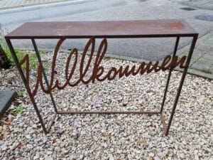 Eine rostfarbene Metallbank mit dem in die Rückenlehne eingearbeiteten Wort „Willkommen“ steht anmutig auf einem Bett aus weißem Kies in der Nähe eines Gehwegs – ein idealer Ort für Paare, die ihre Hochzeitsplanung besprechen.
