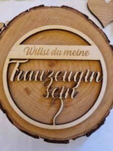 Eine Holztafel in Form einer Baumscheibe mit eingraviertem deutschen Text: „Willst du meine Trauzeugin sein?“ mit Herzausschnitt, perfekt für Ihre Hochzeit. Die Tafel wird auf einer hellen Oberfläche platziert, mit einem kleinen Holzherz in der Ecke – ideal für die Hochzeitsplanung.
