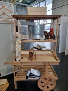 Ein Holzwagen mit kleinem Dach steht drinnen und passt perfekt in jede Hochzeitsumgebung. Er ist mit aufwendigen Holzschnitzereien und runden Schildern mit dekorativer Schrift versehen. Der Wagen verfügt über Holzräder und Regale und ist mit hübschen Holzstücken und Notizbüchern dekoriert, die sich ideal für die Hochzeitsplanung eignen.