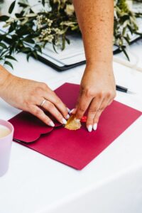 Eine Person versiegelt einen roten Umschlag mit einem goldenen Wachssiegel und gibt damit den Ton für eine elegante Hochzeit an. Daneben bilden eine Tasse und grünes Laub einen ruhigen und dekorativen Hintergrund, perfekt für eine durchdachte Hochzeitsplanung.