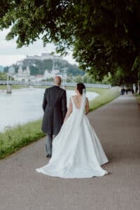 Eine Braut im weißen Kleid und ein Bräutigam im schwarzen Anzug gehen Hand in Hand einen Weg am Flussufer entlang. Bäume säumen den Weg, im Hintergrund sind an diesem verträumten Hochzeitstag eine Brücke und ein Schloss zu sehen.