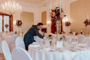 Eine Frau arrangiert eine raffinierte Tischdekoration in einem eleganten Speisesaal, der mit Kronleuchtern und hohen Blumengestecken geschmückt ist. Das anspruchsvolle Ambiente ist perfekt für eine Hochzeit, mit weißen Tischdecken, Tellern und Servietten, die die zarten Blumenarrangements auf jedem Tisch ergänzen.