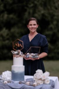 Eine Person in einem dunkelblauen Kleid steht im Freien zwischen Bäumen und hält eine durchsichtige Schachtel mit Keksen in der Hand. Vor ihr, perfekt für jede Hochzeit, steht eine weiß-blaue Torte mit geometrischem Aufsatz und einem Strauß weißer Rosen, elegant auf dem Tisch.