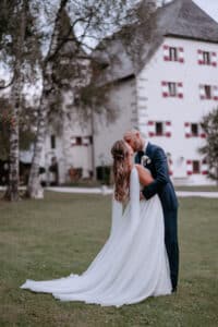 Braut und Bräutigam küssen sich vor einem großen, eleganten Gebäude mit rot-weißen Fensterläden und verkörpern damit den perfekten Moment einer Hochzeit. Die Braut trägt ein fließendes weißes Kleid und der Bräutigam einen dunklen Anzug. Sie stehen auf einer Wiese mit Bäumen im Hintergrund.