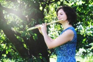 Eine Frau in einem blau gemusterten Kleid spielt Flöte unter einem Baum in einer sonnigen, grünen Umgebung im Freien und verleiht der perfekten Hochzeit eine harmonische Note.