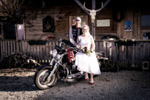 Ein Paar im Hochzeitskleid posiert draußen mit einem Motorrad. Die Braut sitzt anmutig mit ihrem Blumenstrauß in der Hand auf dem Motorrad, während der Bräutigam stolz neben ihr steht. Sie werden von einem rustikalen Holzgebäude umrahmt, das mit bezaubernden Dekorationen geschmückt ist – ein wahrer Beweis für ihre herzliche Hochzeitsfeier.