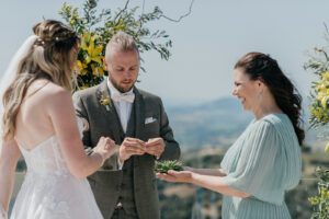 Bei einer bezaubernden Hochzeit im Freien tauschen Braut und Bräutigam die Ringe aus. Der Bräutigam trägt einen grauen Anzug, während die Braut in einem weißen Kleid glänzt. Eine Brautjungfer in Grün hält einen kleinen Blumenstrauß, umrahmt von üppigem Grün und einer malerischen Landschaft, die die Essenz einer makellosen Hochzeitsplanung einfängt.
