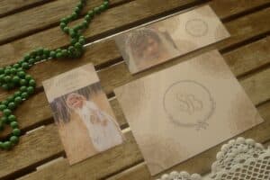 Eine rustikale Hochzeitseinladung auf einem Holztisch fängt die Essenz einer Hochzeit ein. Sie enthält eine Karte mit einem Bild eines Kindes in einem weißen Kleid, den monogrammierten Initialen „SB“, verziert mit Spitzenmustern. Neben den Einladungen liegt eine grüne Perlenkette, perfekt für alle, die schon tief in der Hochzeitsplanung stecken.