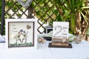 Ein gerahmtes Kunstwerk mit Kieselsteinmenschen und einer herzlichen Karte mit „Yvonne & Hannes 23.07.2022“ ruht auf einem weißen Tisch und fängt die Essenz der Hochzeit ein. Der Hintergrund zeigt ein charmantes Holzgitter, das mit üppigen grünen Pflanzen geschmückt ist.