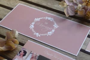 Eine Hochzeitseinladung mit einem rosa Umschlag, auf dem in Weiß die Worte „Die Ehe“ stehen, umrahmt von einem Blumenkranz. Die Eleganz der Hochzeitsplanung wird durch getrocknete Blumen betont, die anmutig daneben auf einer Holzoberfläche platziert sind.