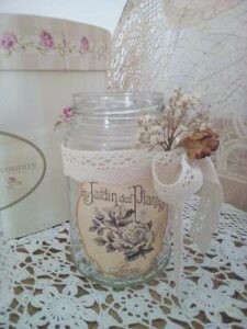 Ein dekoratives Glasgefäß mit einem Vintage-Blumenetikett und Spitzenband, perfekt für die Hochzeitsplanung, ist mit getrockneten Blumen geschmückt. Es steht auf einer mit Spitze bedeckten Oberfläche neben einer Schachtel mit Blumenmuster und verleiht jeder Hochzeitsfeier Charme.