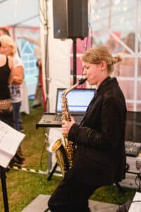 Eine Person im schwarzen Anzug spielt Saxophon in einem Zelt, daneben stehen ein Laptop und Lautsprecher und sorgen so für den perfekten Soundtrack für die Hochzeitsfeierlichkeiten. Im Hintergrund stehen Gäste in Freizeitkleidung und unterhalten sich, was zur fröhlichen Atmosphäre dieses schönen Anlasses beiträgt.