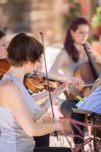 In einer malerischen Umgebung im Freien, die perfekt für eine Hochzeit ist, spielen drei Musiker Streichinstrumente. Der Fokus liegt auf einem Geiger, der in die Musik vertieft ist, während die anderen Musiker leicht verschwommen dahinter zu sehen sind. Sie scheinen die Noten von einem Notenpult abzulesen und verleihen diesem magischen Tag Harmonie.
