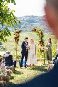 Ein Paar steht während einer Hochzeitszeremonie im Freien vor dem Altar, vor der Kulisse sanfter Hügel und Weinberge. Der Altar ist mit bunten Blumen geschmückt. Die Gäste sitzen auf Heuballen, was eine rustikale Hochzeitsplanung verkörpert, während eine Frau in der Nähe mit einem Notenständer steht.