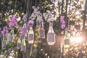 Hängende, mit Schnur umwickelte Glasflaschen dienen als bezaubernde Vasen für rosa und weiße Blumen und verleihen der Hochzeitsdekoration eine romantische Note. Sie baumeln an Seilen vor einem Hintergrund aus üppigem Grün und gesprenkeltem Sonnenlicht, perfekt für den besonderen Tag der Hochzeit des Paares.