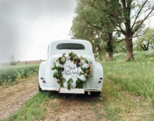 Ein Oldtimer mit einem Blumenkranz und einem „Just Married“-Schild auf der Rückseite steht auf einer Landstraße, perfekt für alle, die ihre Traumhochzeit planen. Üppiges Grün und hohe Bäume säumen die Straße und schaffen eine ruhige und romantische Atmosphäre.