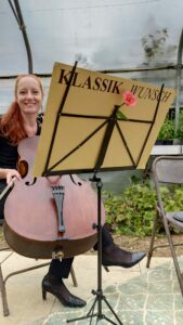 Eine Frau mit langen roten Haaren sitzt auf einem Stuhl und hält ein Cello in der Hand. Vor ihr steht ein Notenständer mit einem Blatt mit dem Titel „Klassik Wunsch“ und einer daran befestigten rosa Blume. Sie steht unter einem transparenten Baldachin in einem Garten – eine ruhige Kulisse, perfekt für jede Hochzeitsfeier.