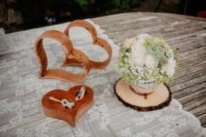 Eine rustikale Hochzeitsszene zeigt zwei ineinander verschlungene Herzdekorationen aus Holz, eine kleine herzförmige Holzschachtel mit zwei Ringen und einer Schleife sowie einen Blumenstrauß in einer mit Sackleinen umwickelten Vase auf einem Holztisch mit Spitzenauflage.