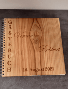 Ein hölzernes Gästebuch mit elegant vertikal eingraviertem „Gästebuch“ auf der linken Seite, perfekt für Ihre Hochzeit. Die Namen „Verena & Robbert“ und das Datum „14. August 2021“ sind in einer anmutigen Schreibschrift eingraviert und halten unvergessliche Momente Ihres besonderen Hochzeitstages fest.