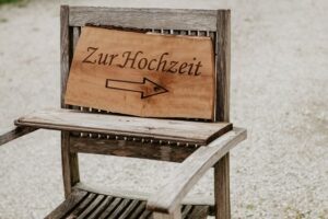 Auf einem Holzstuhl steht ein Schild mit der Aufschrift „Zur Hochzeit“ und einem nach rechts zeigenden Pfeil, der den Gästen den Weg zur Hochzeit weist. Perfekt für alle, die schon mitten in der Hochzeitsplanung stecken, bietet die Außenanlage einen charmanten Kiesweg, der den Weg weist.