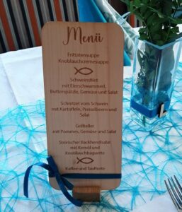 Eine vertikale Holzkarte auf einem Tisch, geschmückt mit einer blauen Schleife und begleitet von einer blauen Vase, sorgt für eine gemütliche Hochzeitsatmosphäre. Die Auswahl umfasst Frittatensuppe, Schweinsfilet mit Eierschwammerl, Schnitzel vom Schwein, Grillteller, Steirischer Backhendlsalat und Kaffee und Tauforte. Perfekt für Ihre Hochzeitsplanung